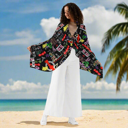 Akihabara Pulse Wrap Sarong displayed on a tropical beach setting, featuring vibrant neon-inspired gaming patterns in red, green, and white against a black background. The flowing sarong showcases wide kimono-style sleeves and contrasts beautifully with white pants and the turquoise ocean backdrop.