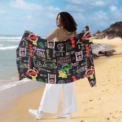 Akihabara Pulse Wrap Sarong displayed on beach, featuring black fabric with colorful Japanese-inspired neon signs and symbols pattern, held up against ocean backdrop with blue sky and sandy shore, showcasing its generous size and vibrant design