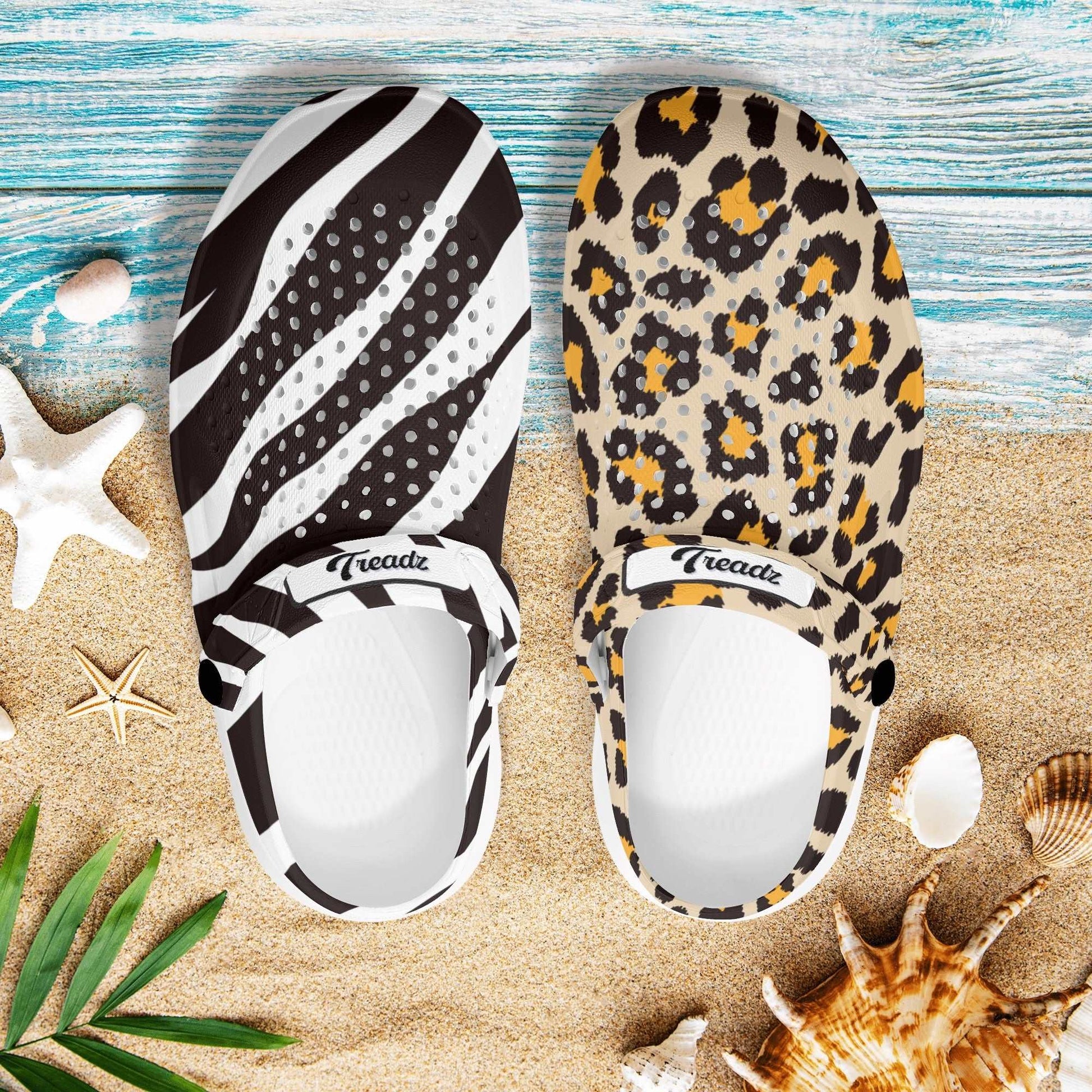 Treadz Mismatch Clogs - Safari displayed on sandy beach with one clog featuring zebra stripes and the other leopard print, shown with seashells and tropical elements on turquoise wooden background