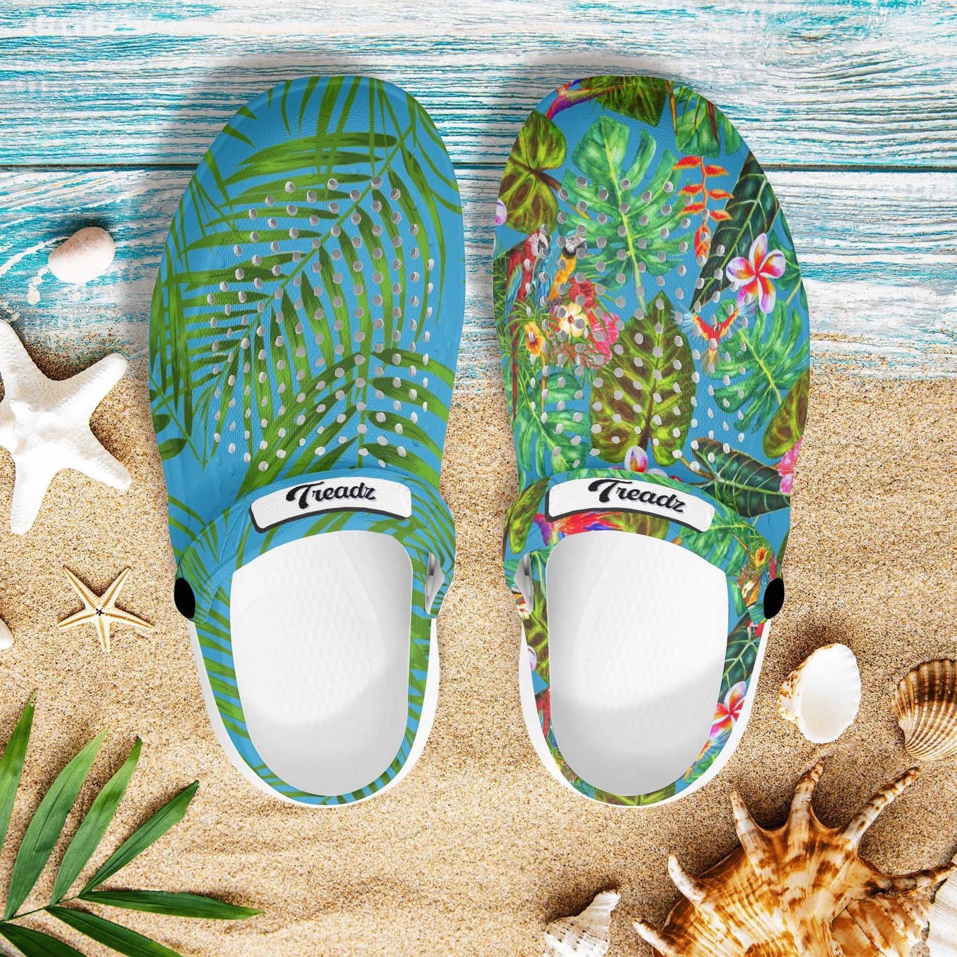 Treadz Mismatch Clogs - Tropicana: Mismatched EVA clogs featuring tropical designs - one with green palm leaves on blue, the other with colorful birds and foliage, displayed on beach-themed background with seashells and starfish