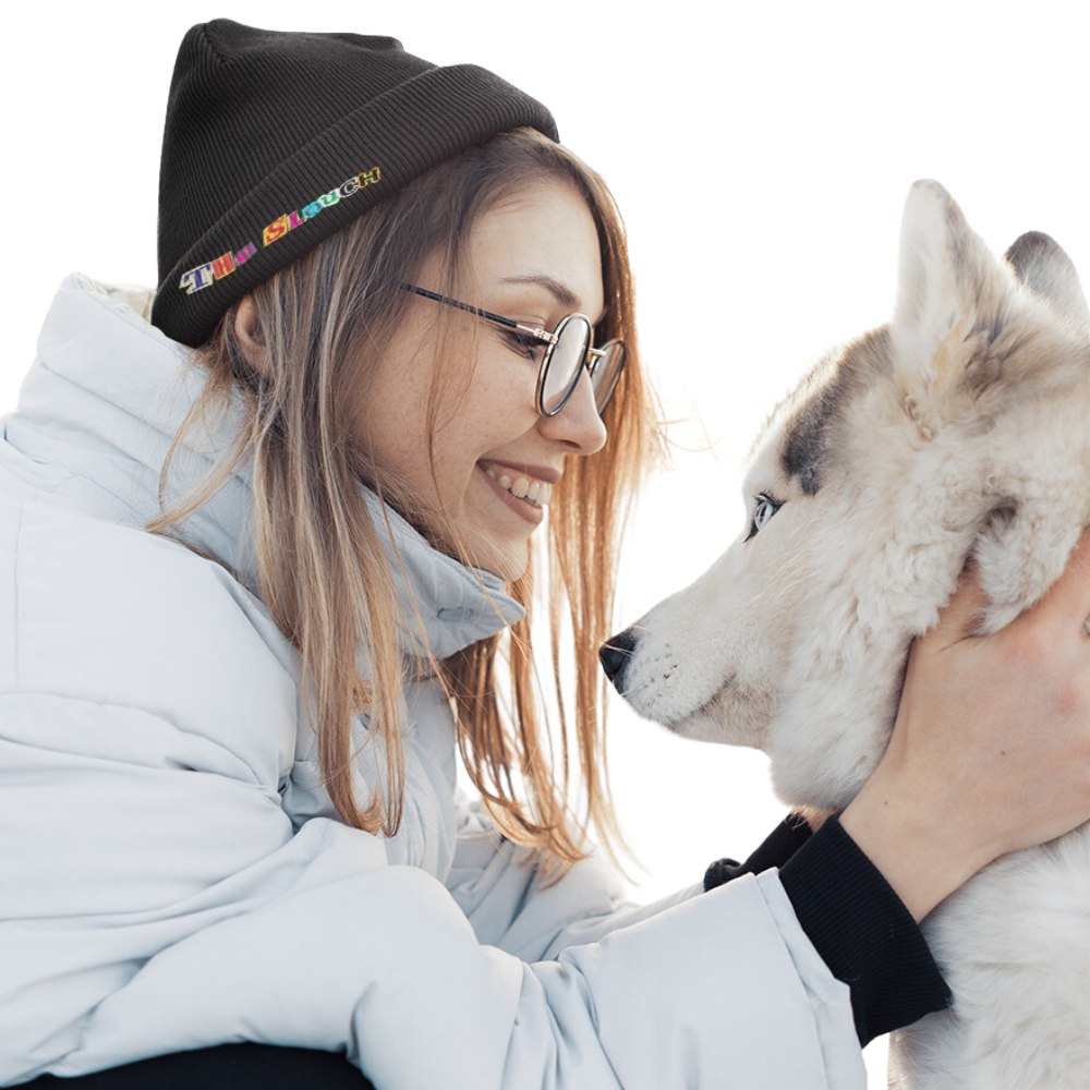 Black Slouch Beanie shown in a winter scene with a white husky dog, featuring a small colorful logo detail on the beanie. The wearer is dressed in a light-colored winter jacket, creating a cozy outdoor winter atmosphere.