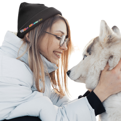 Black Slouch Beanie shown in a winter scene with a white husky dog, featuring a small colorful logo detail on the beanie. The wearer is dressed in a light-colored winter jacket, creating a cozy outdoor winter atmosphere.