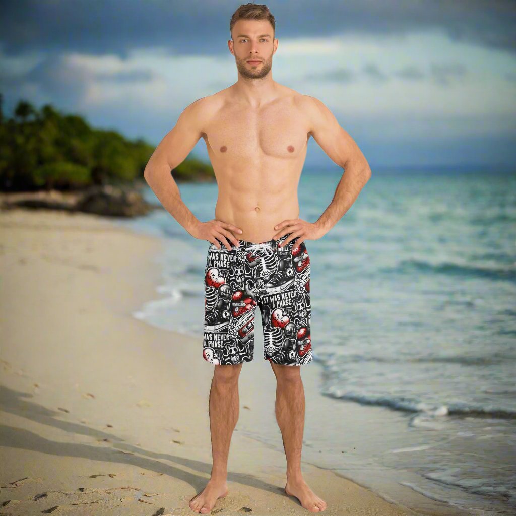 Rip Tide Men's Board Shorts - Emo Vibes shown on a beach backdrop, featuring black and white skeleton print with red accents. Model wearing the shorts stands on sandy beach with ocean waves and palm trees in background.