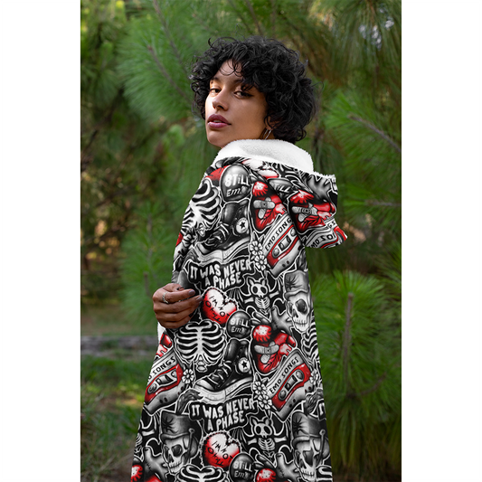 Womens Hooded Blanket - Emo Vibes featuring edgy black, white and red skull pattern design displayed in outdoor setting with pine trees. Cozy fleece blanket with hood shown draped over shoulders, highlighting the all-over gothic print and plush material. ShitHot Best Sellers Collection.